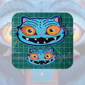 May include: Two embroidered patches featuring a cartoon cat face with a blue body, orange eyes, and a black mouth. The patches are on a green cutting mat. The larger patch is approximately 4 inches wide.