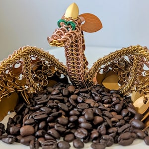 Peut inclure: Une sculpture de dragon artisanale en cotte de mailles de cuivre et de laiton, avec une tête et des ailes dorées. Le dragon est perché au-dessus d'un tas de grains de café torréfiés foncés, créant une pièce décorative unique.