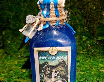 Könnte beinhalten: Eine kleine blaue Glasflasche mit einem durchsichtigen Kristallverschluss. Die Flasche ist mit Schnur und blauen Wachstropfen verziert. Das Etikett lautet "Siren's Song" und zeigt eine Illustration einer Meerjungfrau.