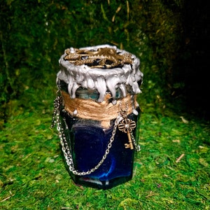 May include: A small blue glass jar with a silver and gold lid, decorated with a chain and a key. The jar is sitting on a bed of green moss.