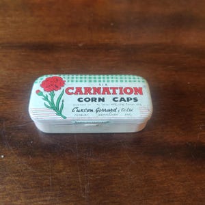 Op de afbeelding: Een vintage metalen blik voor Carnation Corn Caps. Het blik heeft een rode anjerbloemillustratie en tekst. De tekst bevat "CORN CAPS" en de naam van de fabrikant. Het blik is rechthoekig met afgeronde randen en een gestreept patroon.