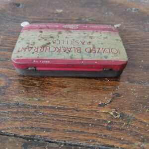May include: Vintage rectangular tin with a red and cream color scheme. The tin reads "IODIZED BLACKCURRANT PASTILLES" in red lettering. The tin is slightly rusted and sits on a wooden surface.