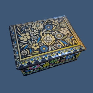 Può includere: Una scatola rettangolare in metallo con base blu scuro e un vivace motivo floreale nei toni del giallo, azzurro, bianco e verde. Il coperchio e i lati sono decorati con complessi motivi di fiori e foglie, creando un design denso e colorato sull'intera superficie esterna del contenitore.