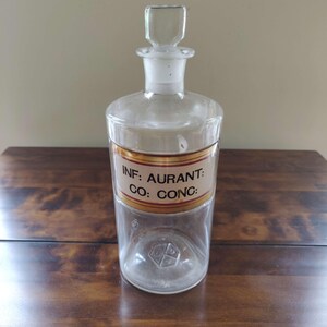 May include: A clear glass apothecary bottle with a stopper. The bottle has a label with the text "INF: AURANT: CO: CONC:" in black lettering. The label is bordered by a gold and brown band. The bottle is on a wooden surface.