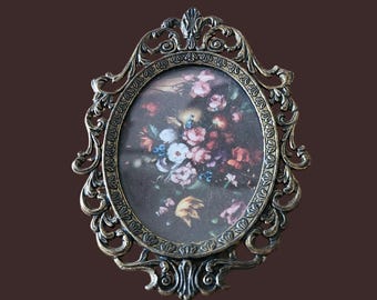 Könnte beinhalten: Ovale Bilderrahmen mit aufwendigen Verzierungen und Bronze-Finish. Der Rahmen umschließt einen Blumendruck mit rosa, weißen und blauen Blumen. Ein dekoratives Element für die Inneneinrichtung, ideal für ein Foto.