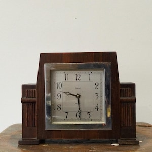 Puede incluir: Un reloj de madera vintage con una esfera cuadrada y un borde plateado. La esfera del reloj muestra los números del 1 al 12 y las manecillas señalan aproximadamente las 9:30. El reloj es de madera marrón oscuro y tiene forma rectangular.