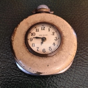 May include: A vintage pocket watch featuring a cream-coloured case and a circular clock face. The clock face displays black numerals and hands, with the words "NEW HAVEN" printed below the 12. The watch has a silver-coloured winding stem.