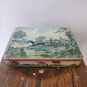 Puede incluir: Caja de lata rectangular con una escena de caza pintada. La tapa presenta jinetes a caballo, perros y un paisaje con árboles y una valla. La caja tiene un borde rojo y una escena similar en el lateral.