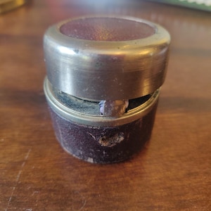 Puede incluir: Un recipiente vintage de dos piezas con exterior de cuero burdeos oscuro y borde de metal color latón. La parte superior e inferior son circulares, siendo la superior ligeramente más pequeña. El recipiente muestra signos de desgaste.