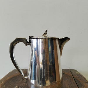 Op de afbeelding: Een zilverkleurige kan met een gebogen handvat en een puntige tuit. De kan heeft een deksel met een kleine, decoratieve knop. Het oppervlak van de kan is zeer reflecterend en toont de omgeving. De kan is waarschijnlijk van metaal en staat op een houten oppervlak.