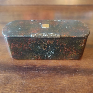 Puede incluir: Una caja rectangular antigua con un acabado moteado negro y rojo. La caja tiene un cierre dorado y adornos decorativos en el borde superior. La caja está sobre una superficie de madera.