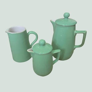 Può includere: Servizio da tè in ceramica verde menta composto da tre pezzi. Il set include una lattiera, una piccola teiera con coperchio e una teiera più grande con coperchio. L'interno della lattiera è bianco. Le teiere hanno una forma arrotondata con manico e beccuccio.