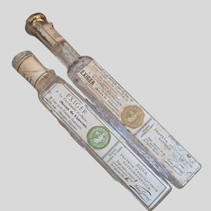May include: Two antique glass bottles with labels. The labels feature text in French and Latin, along with decorative seals. The bottles have gold-colored caps and are rectangular in shape.