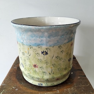 May include: A ceramic planter featuring a hand-painted landscape design. The upper portion displays a light blue sky, while the lower section depicts a green meadow filled with small, scattered wildflowers in shades of red, yellow, and blue.