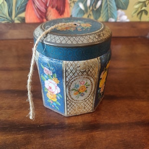 Puede incluir: Lata hexagonal vintage con diseño floral. La lata presenta un fondo azul con detalles en blanco y dorado, y un asa de cuerda. La tapa y los lados están decorados con flores coloridas.