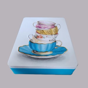 May include: A rectangular, light blue box with a white top featuring a watercolor illustration of stacked teacups. The teacups are in shades of pink, yellow, white, and blue, with gold accents. The bottom teacup sits on a matching blue saucer.