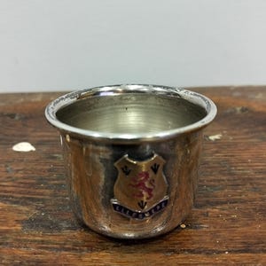 May include: A small, silver-colored cup with a slightly worn rim. The cup features a crest with a red lion and the word "ELLSMERE" in blue lettering. The cup sits on a wooden surface.