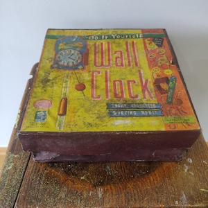 Pode incluir: Uma caixa de kit de relógio de parede "Do It Yourself" vintage. A caixa tem um fundo amarelo com um gráfico de relógio e as palavras "Wall Clock" em letras vermelhas grandes. A caixa está sobre uma superfície de madeira.