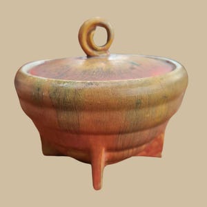 May include: A small, round, ceramic pot with a lid. The pot is decorated with a brown, orange, and green glaze. The lid has a small, looped handle. The pot sits on three short legs.