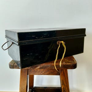 May include: A black metal box with a key in the lock, resting on a small wooden stool. The box has a handle on the side and looks vintage. The key has a yellow ribbon attached. The stool is made of wood with a natural finish.
