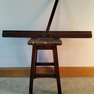 Könnte beinhalten: Ein altes Zeichenwerkzeug aus Holz mit einem langen Lineal mit Messmarkierungen steht auf einem kleinen, alten Holzschemel. Das Werkzeug hat einen rechtwinkligen Arm. Der Hocker hat eine quadratische Sitzfläche und vier Beine.