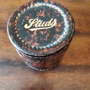 Könnte beinhalten: Ein zylindrischer Behälter mit Schildpattmuster und dem Wort "Studs" in goldfarbenen Buchstaben auf dem Deckel. Der Behälter besteht aus einem dunkelbraunen Material und hat einen genähten Rand.