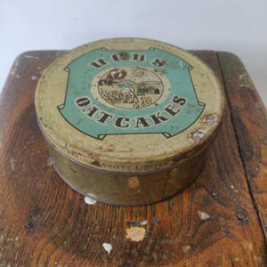 Puede incluir: Una lata redonda de metal vintage, en tonos dorados y verde claro. La lata presenta la palabra "Oatcakes" en una fuente curva, con una ilustración central de una persona en un campo. La lata muestra signos de óxido y desgaste.