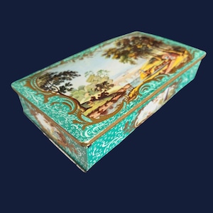 Puede incluir: Caja rectangular turquesa con una escena de paisaje en la tapa. La caja presenta adornos dorados y una pintura detallada de paisaje con árboles, figuras y cielo azul. Los lados de la caja tienen un patrón floral blanco.