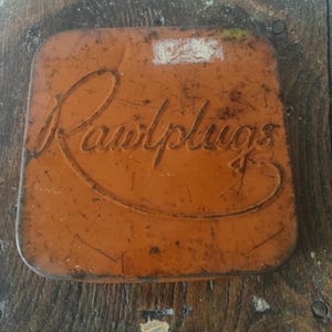 May include: An orange, square metal container with the word "Rawlplugs" embossed on the lid. The container has a distressed, aged appearance, with signs of wear and rust. The background is a wooden surface.