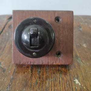 Puede incluir: Un interruptor de luz vintage con un interruptor basculante circular marrón oscuro montado sobre una base de madera rectangular. El interruptor está asegurado con tornillos y tiene un diseño clásico y retro. La base de madera tiene un tono cálido.