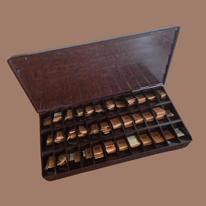 May include: A dark brown plastic storage case with a hinged lid, containing multiple compartments filled with small, light tan rectangular cards. Each card features printed black text, including various letter combinations like aa, oo, and eu.