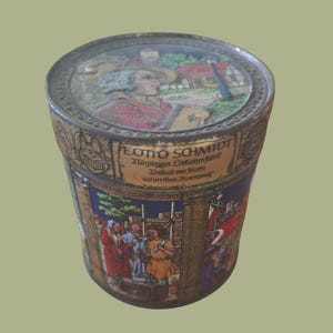 May include: Vintage cylindrical tin with a detailed illustration. The lid features a portrait of a man in a hat. The body of the tin displays a scene with figures in period clothing, and text in German. The tin is primarily gold, blue, and red.
