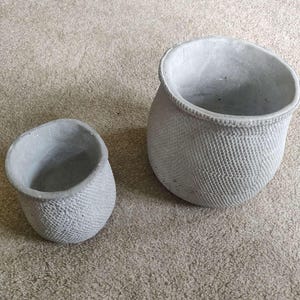 May include: Two grey concrete planters with a textured surface. The larger planter is taller and wider than the smaller planter.