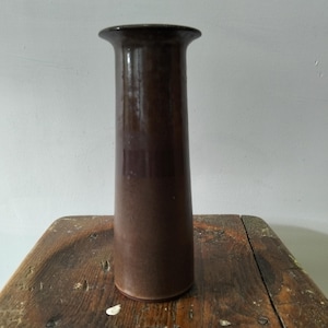 May include: A tall, cylindrical ceramic vase with a smooth, dark brown glazed finish. The vase features a slightly flared, wide rim at the top and a straight, uniform body that tapers very subtly towards the base, creating a sleek profile.