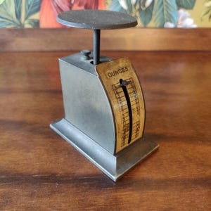 Puede incluir: Una báscula de metal vintage con una plataforma circular y una cara curva que muestra "OUNCES". La báscula es gris con una apariencia oscura y envejecida, lo que sugiere que es una antigüedad. La báscula está diseñada para medir en onzas.