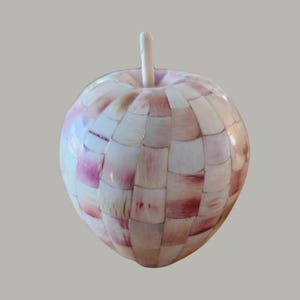 Può includere: Una mela a mosaico rosa e bianca realizzata con pezzi di conchiglia. La mela ha una forma rotonda e liscia e un piccolo gambo in cima.