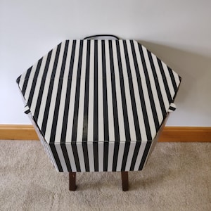 May include: A black and white striped hexagonal storage box with a black handle. The box is supported by four small, dark brown wooden legs. The box is made of cardboard or a similar material.