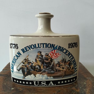 May include: A cream-coloured ceramic decanter featuring a graphic of the American Revolution. The text reads 1776, 1976, AMERICAN REVOLUTION BICENTENNIAL, and U.S.A. with stars. The illustration depicts figures in a boat on water with a flag.