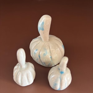 May include: Three ceramic pumpkin figurines with a glossy off-white finish and subtle light blue speckles. Each pumpkin features a rounded body with vertical ridges and a smooth, upright stem. The set includes one larger and two smaller pieces.