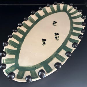 May include: Oval ceramic serving platter with a cream-coloured centre and a green striped border. The rim is adorned with evenly spaced, glossy black spheres. Small black floral designs are scattered across the centre.