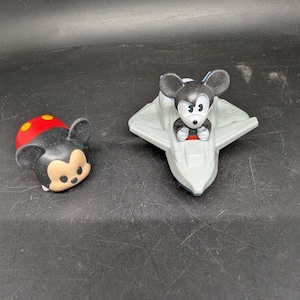 May include: Two Disney Mickey Mouse figurines. One is a Tsum Tsum style figure with a red and yellow top and black face. The other is Mickey Mouse in a gray spaceship. Both are on a dark surface.