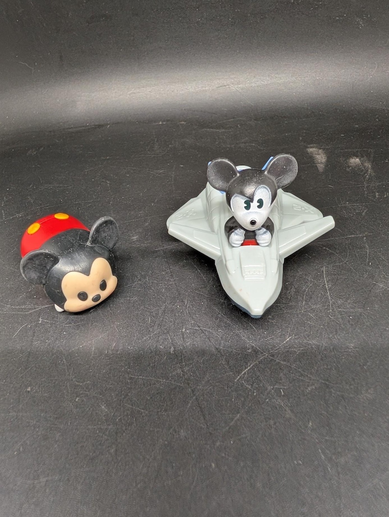 May include: Two Disney Mickey Mouse figurines. One is a Tsum Tsum style figure with a red and yellow top and black face. The other is Mickey Mouse in a gray spaceship. Both are on a dark surface.