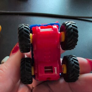 May include: A small, red toy truck with black tires and yellow wheel hubs. The truck has a blue top and is viewed from underneath. The tires have a deep tread pattern. The text "1934AZ" is visible on the truck.