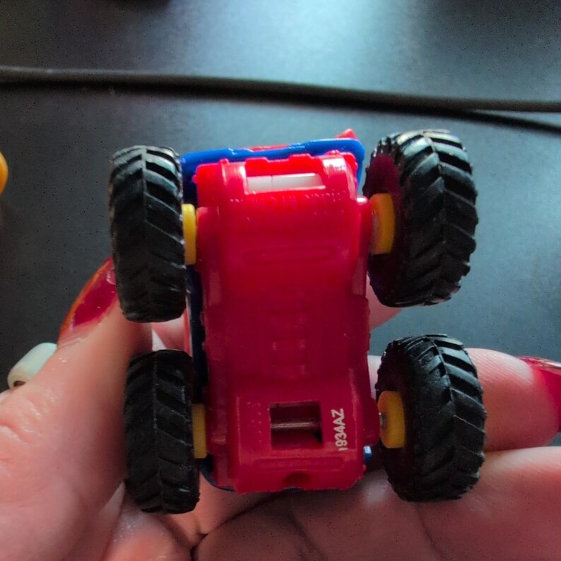 May include: A small, red toy truck with black tires and yellow wheel hubs. The truck has a blue top and is viewed from underneath. The tires have a deep tread pattern. The text "1934AZ" is visible on the truck.