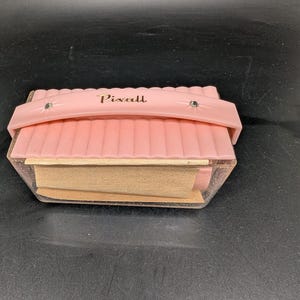 Könnte beinhalten: Ein vintage rosa Pixall Nähset mit einer transparenten, glitzernden Kunststoffbasis. Der Deckel hat einen Griff und ist mit zwei silberfarbenen Nieten befestigt. Das Wort "Pixall" ist in goldfarbenen Buchstaben auf den Deckel gedruckt.