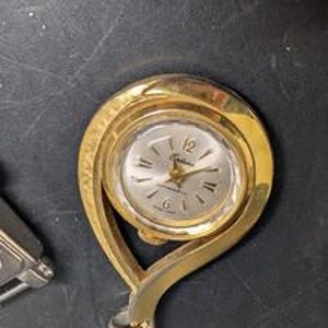 May include: A gold-toned vintage pendant watch with a silver-colored face. The watch has a curved, open design and a small loop at the bottom. The watch face displays the numbers 12, 3, 6, and 9.