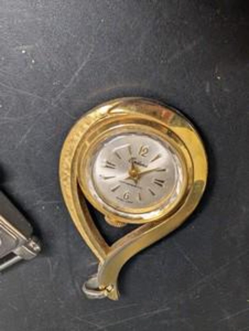 May include: A gold-toned vintage pendant watch with a silver-colored face. The watch has a curved, open design and a small loop at the bottom. The watch face displays the numbers 12, 3, 6, and 9.
