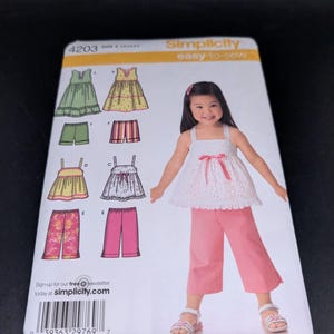 May include: A Simplicity 4203 easy-to-sew pattern envelope featuring illustrations of various children's clothing designs including dresses, shorts, and pants. The cover displays a child wearing a white eyelet top with a pink bow and pink pants.