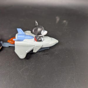 May include: A gray toy rocket with blue wings and orange flames. A black and white dog figurine is seated in the cockpit. The toy is on a dark surface.