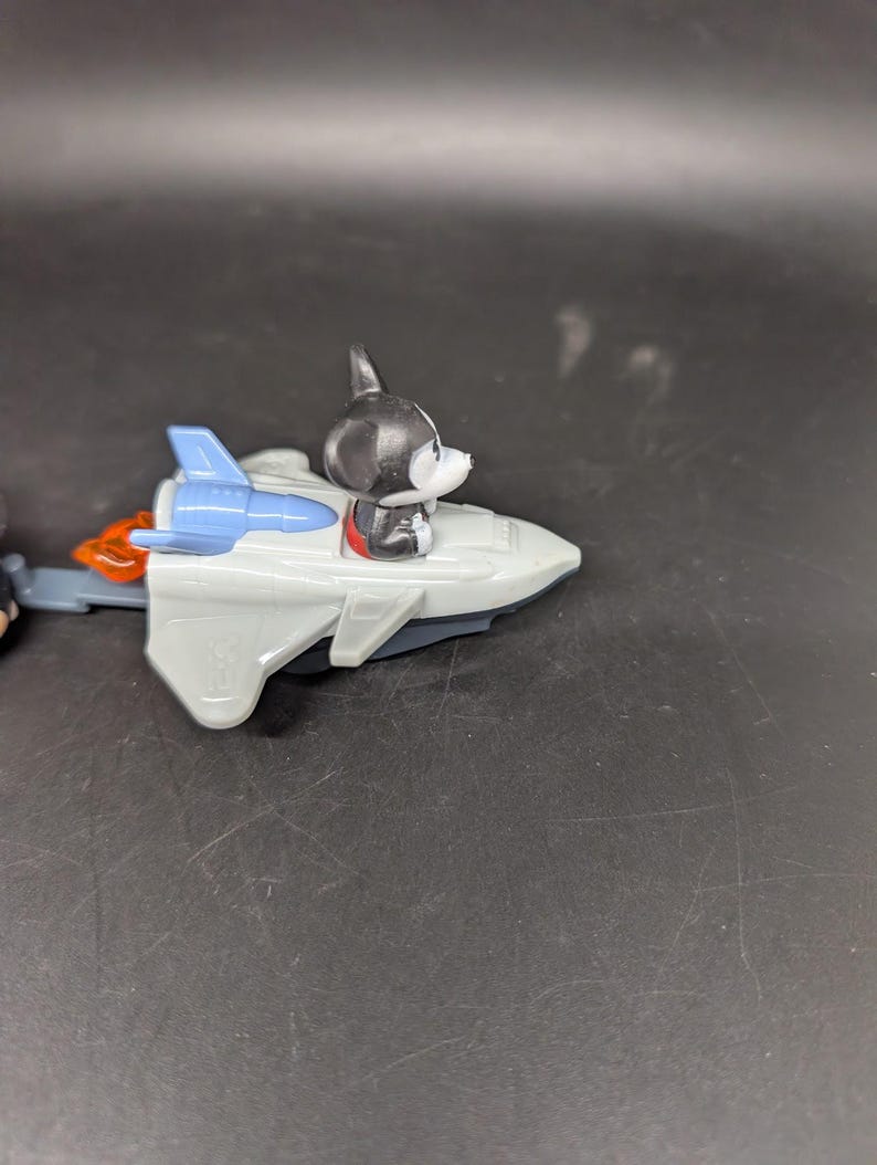 May include: A gray toy rocket with blue wings and orange flames. A black and white dog figurine is seated in the cockpit. The toy is on a dark surface.